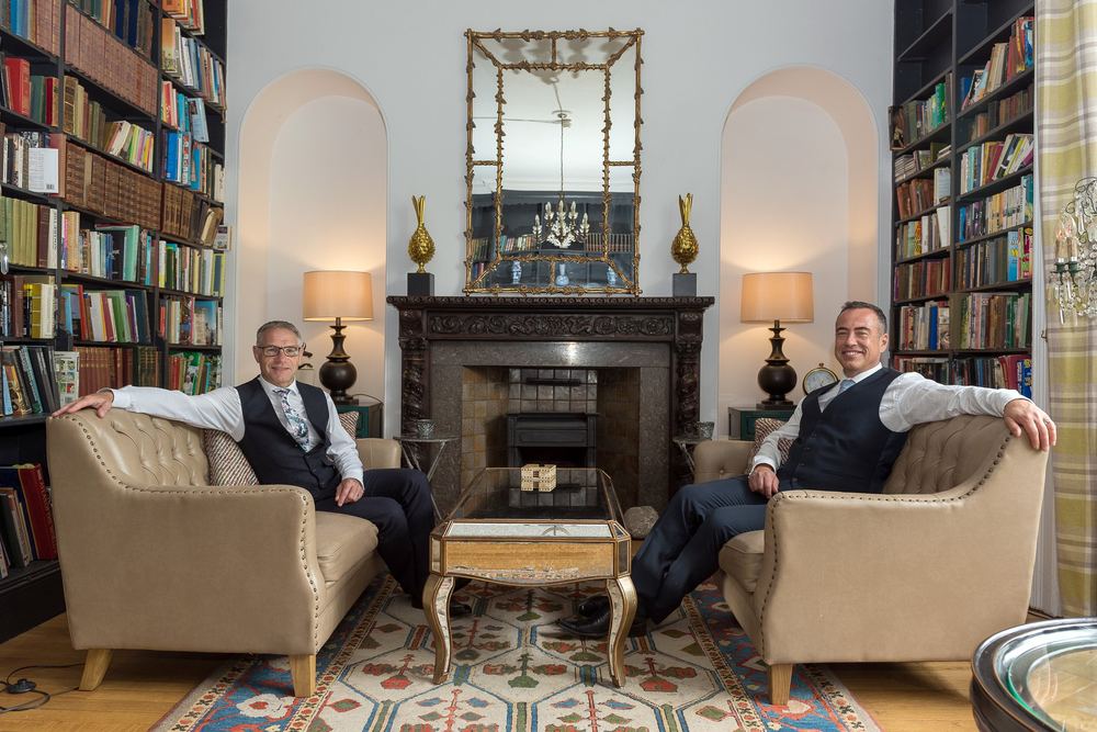 Two men in matching waistcoats, seated either side of a library fireplace.