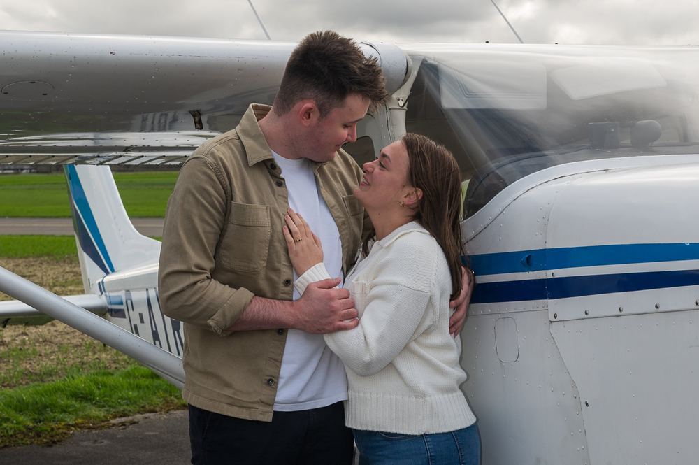 A newly engaged couple look adoringly at each other beside their plane.