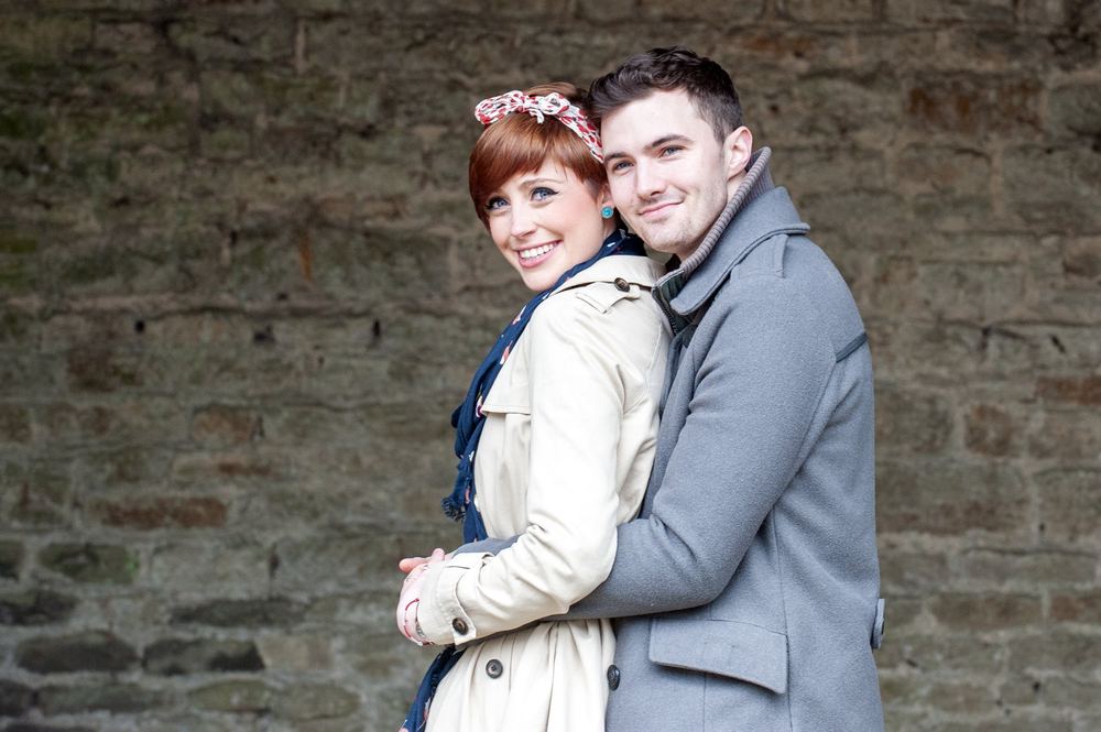 A young couple cuddle in front of an old brick wall.